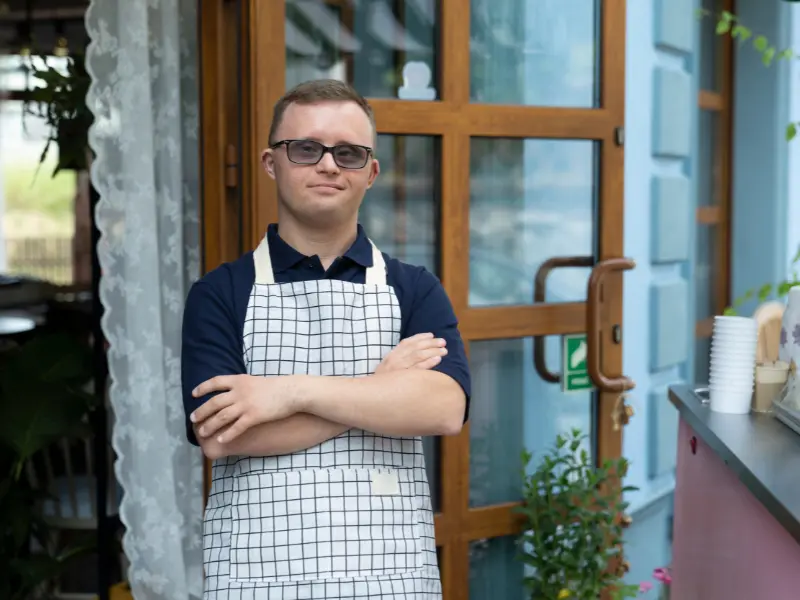 Disabled man with down syndrome smiling at camera with arms crossed wearing an apron at work