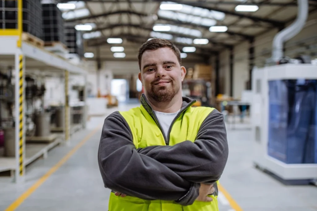 Disabled man with down syndrome in warehouse with high vis vest on smiling at camera with arms crossed