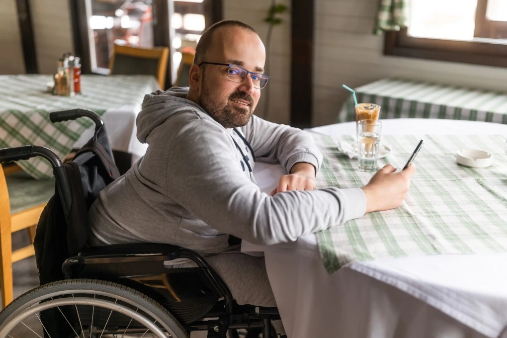 Quadriplegic man sitting at cafe using mobile phone and smiling at camera